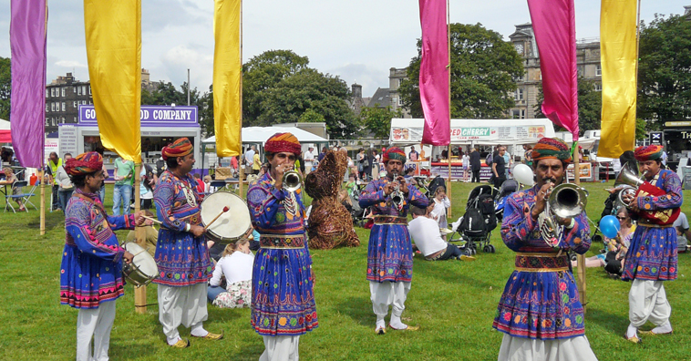 Men in purple tunics with white trousers and red and orange turbans playing brass instruments with yellow and pink penants in the background