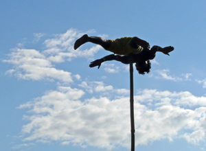 The man pictured left (on the right) spins on his stomach atop a  high pole with clouds in the background