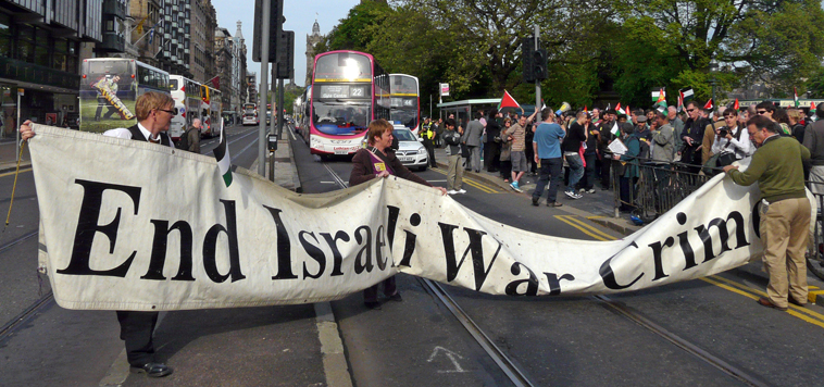 "End Israeli War Crimes" banner being organised  with buses behind, while man with loud hailer explains what is happening