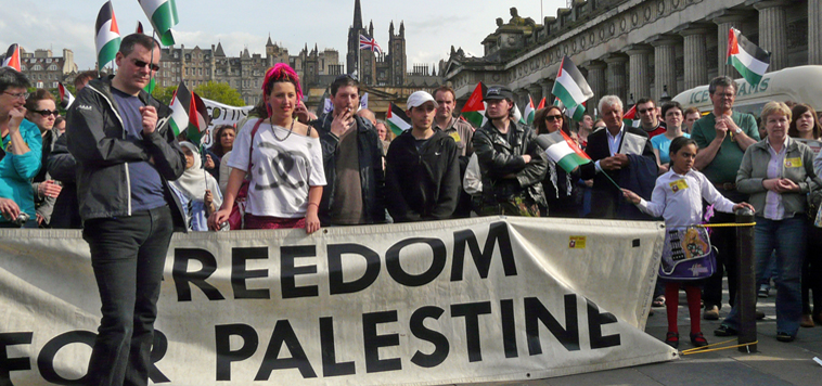 People lined behind a banner "Freedom for Palestine" , waving Palestinian flags. The  National Galleries Mound Complex is in the background