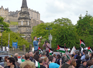 Man with loud hailer surrounded by people waving Palestinian flags with Edinburgh Castle in the background