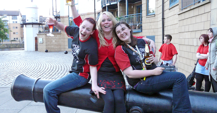 Hazel and Sarah of Pulse of the Place in red and black seated on a cannon, with Louise Marshall Millington (centre) raising their glasses after an evening's musical performance