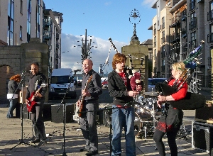 Two guitarists and two pipers performing with Leith Docks in the distance