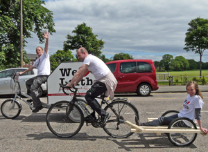 Two men on bicycles , pulling trailers - one with a young woman seated, the other  with the We Heart Leith logo
