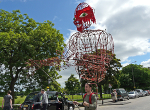 Man walking past with a  seven foot high red faced wicker puppet on his shoulders