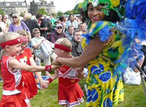 Three girls in red and white cheer leader's costumes being encouraged to join in the dancing by woman in Brazilian flag coloured costume