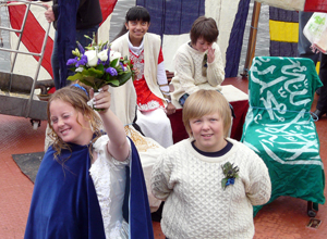 Courtney (in white gown, with blue cloak,) raises a purple and white bouquet as  Lewis (in wbite fisherman's sweater with kilt,) looks up at the waitng crowd