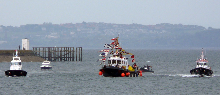 Boat with many coloured penants flanked by pilot boats and two smaller boats approaching with  the  Kingdom of Fire in the background