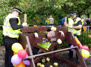 Two Police Officers operating the stocks as another has yellow sponges thrown at his head