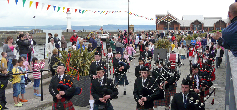 Boys Brigade Piper Band leading procession down harbour wall with Newhaven lighthouse in the background