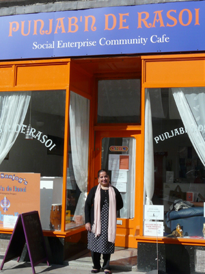 Woman standing out side the orange and blue frontage with the sign "PUNJAB'N DE RASOI Social Enterprise Community Cafe