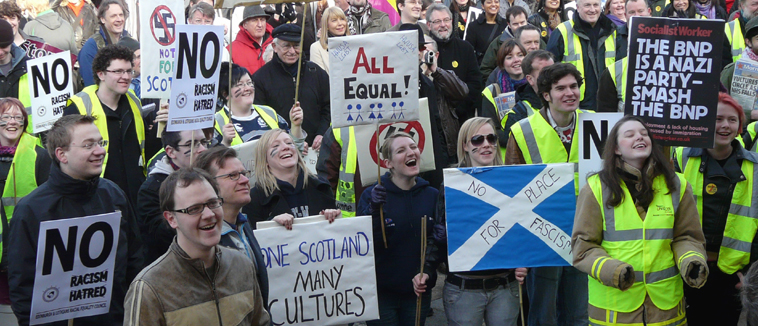 Students laughing at a joke by Weyman Bennett and holding home made placards with slogans including "ONE SCOTLAND MANY CULTURES"  and "NO PLACE FOR FASCISM" on a Saltire