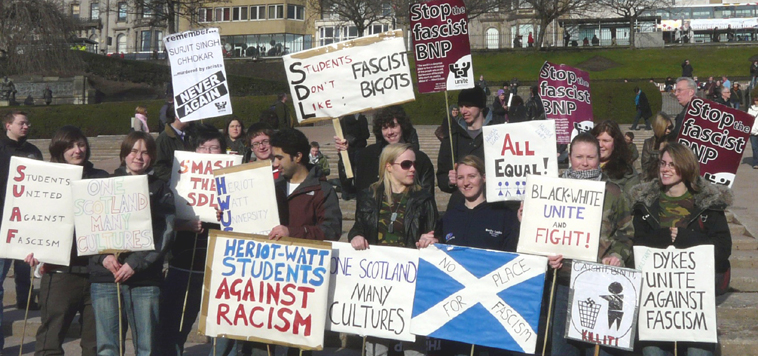 Students posing with home made placards with slogans including "STUDENTS DON'T LIKE FASCIST BIGOTS" and "DYKES UNITE AGAINST FASCISM" and "CATCH IT, BIN IT, KILL IT"