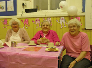 Three ladies in pink enjoying tea and bacon rolls with pink figures on the wall behind them