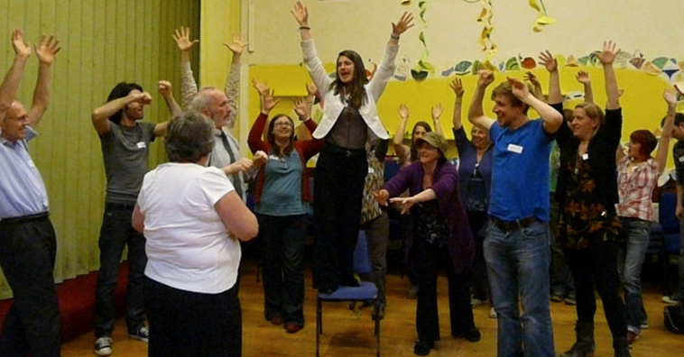 Woman in white jacket and black jeans stands on chair leading group of standing people to raise their arms