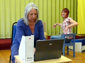 Woman seated in powder blue cardigan, hunched over her laptop as a woman seated behind her talks to her