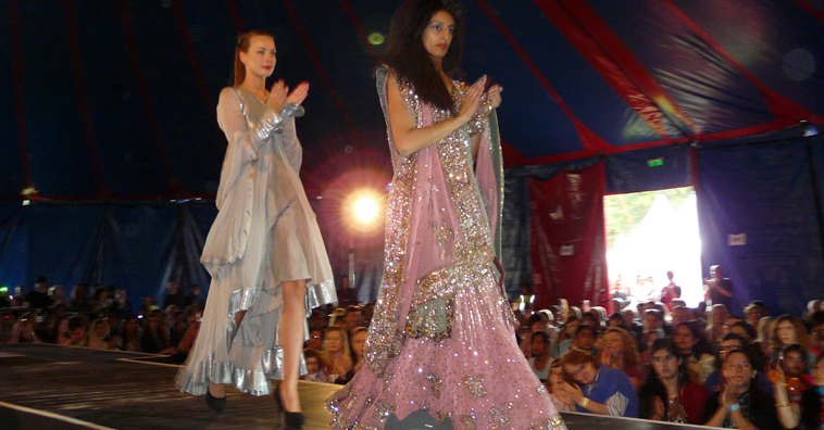 Two women walking along a catwalk, applauding - one in sliver evening gown, one is pink evening gown with glittering embroidery