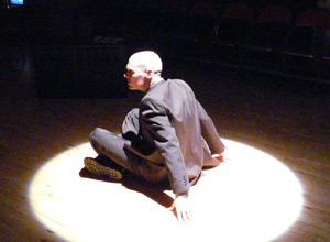 Man in a grey pin striped suit, seated on the ground under a bright spotlight