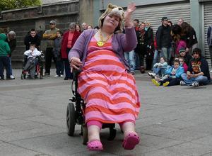 Woman in a pink and red striped dress and a Lion mask dancing in a wheelchair as people smile and applaud
