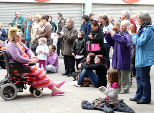 Jessie in wheelchair, Lion King mask, and red and pink striped dress emoting with her hand