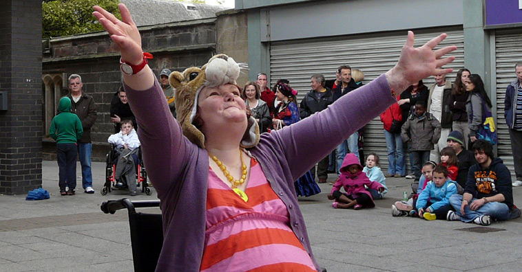 Woman in a pink and red striped dress and a Lion mask dancing in a wheelchair looks to the sky with arms outstretched