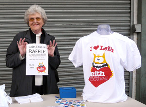 Woman holding up a flyer at a stall covered in buttons with a I heart Leith tee shirt