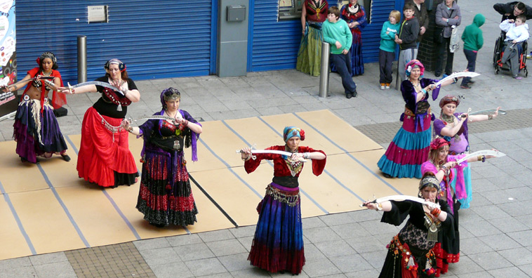 View of a half dozen members of Twisted Tails raising their swords in front of their faces, surrounded by an audience in the Kirkgate