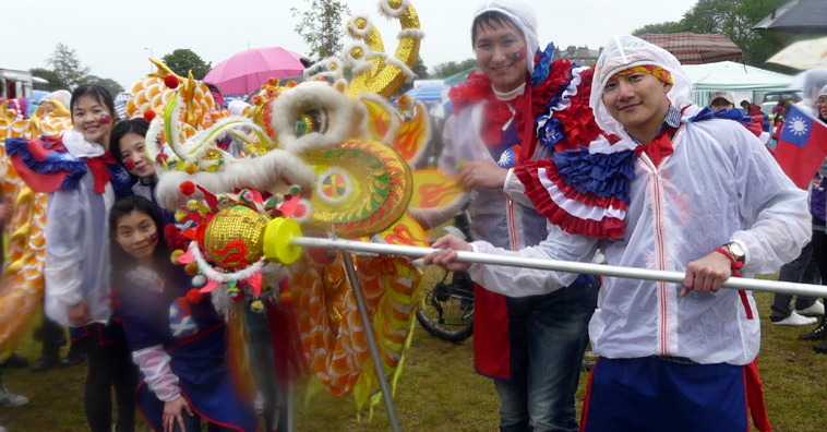 Five students in blue and red, with white waterproof ponchos, pose with a white and gold dragon's head
