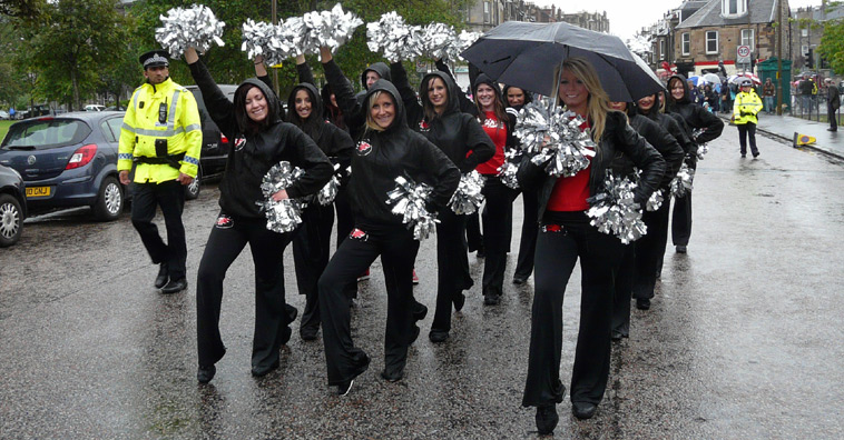 Women in black costumes with hoods strile a pise with silver pom poms