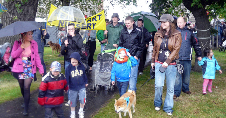 Parents and small children and a huskie with umbrellas and a yellow banner HEADSTART NURSERY passing underneath trees