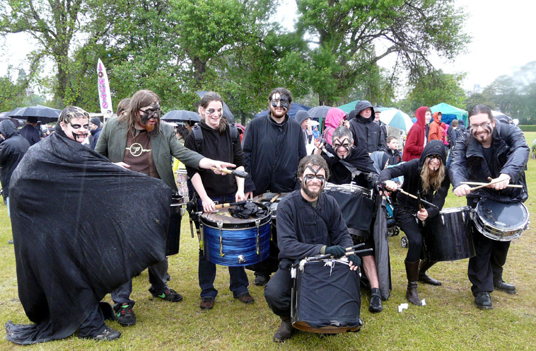An eight piece drumming band all in black with facepaint in a group pose