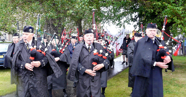 Black clad pipers in waterproofs with red trimmed pipes marching on to Leith Links