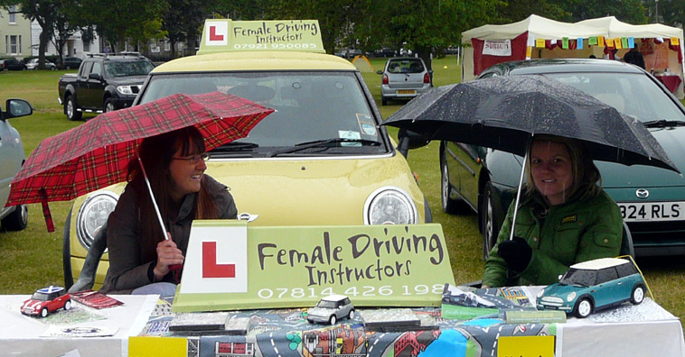 Two women sitting beneath umbrellas at the Female Driving Instructors stall