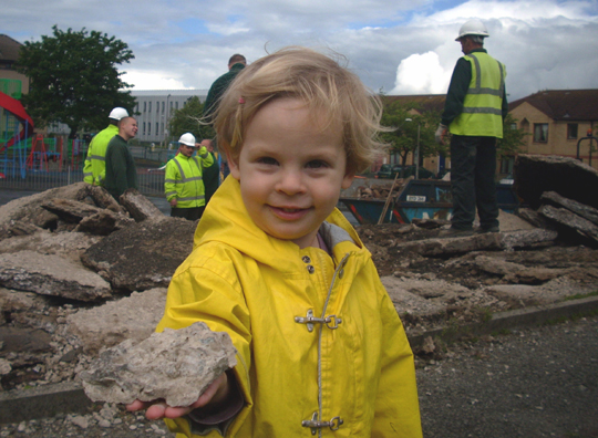 Little girl in yellow waterproof holding out piece of rubble, as men break up concrete behind her.