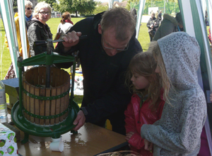 Man pouring juice from a small open topped barrel with green metal press, as two girls watch on