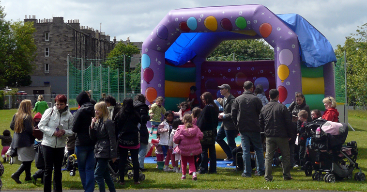 Purple bouncy castle with coloured balloons stamped on it, surrounded by parents and small children