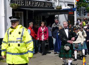 Uniformed officer in yellow waterproofs followed by piper and the Queen's Party