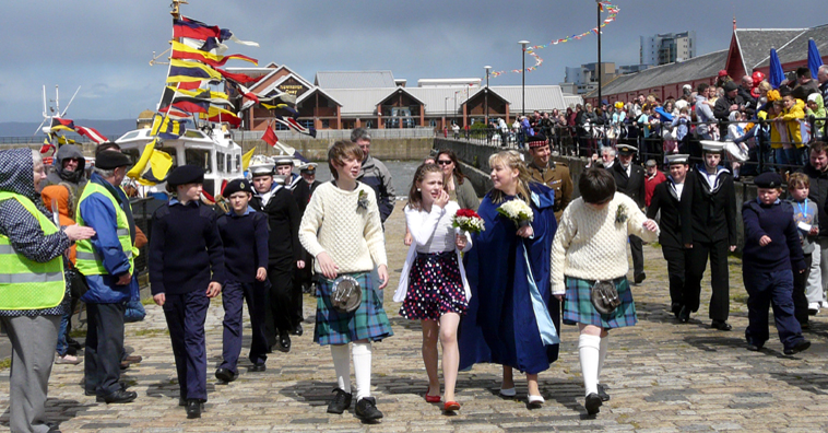 Four children boys in fishermans's sweaters and kilts, the Queen in purple gown with Sea Cadets to either side