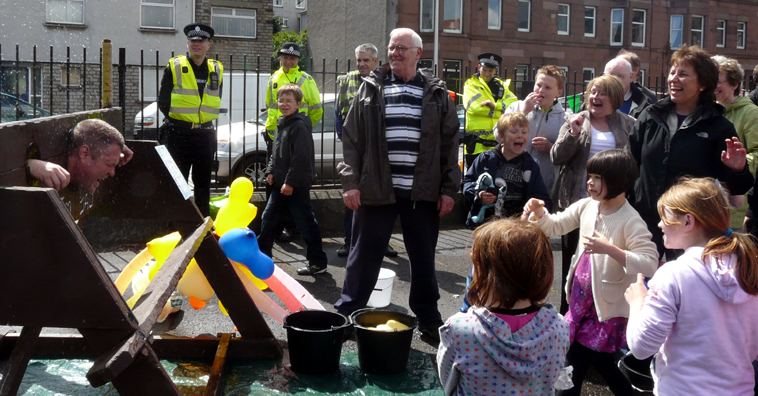Man in the stocks with his head covered in water from a wet sponge thrown by a little girl, surrounded by people laughing
