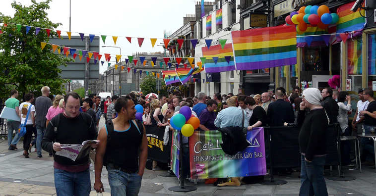 Two men walking past a crowded Cafe Habana decled with rainbow bunting, flags and balloons