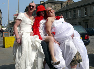 Two shaven headed men in white bridal dresses and a woman in white hitching their skirts