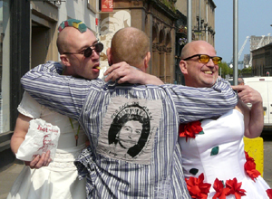 Back view of a man with a striped Sex Pistols tee shirt hugging two men in bridal gowns round their necks