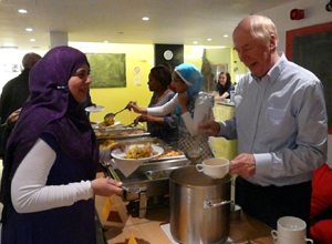Malcolm serving soup to a woman in a purple head scarf